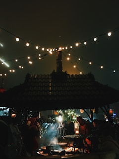 A cozy outdoor setting with a group of people gathered around a fire pit under a wooden shelter. String lights hang above, illuminating the scene with a warm glow. Smoke rises from the fire, adding to the ambiance. In the background, there are festive decorations and greenery, suggesting a holiday or winter market setting.