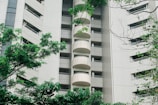 A high-rise apartment building with a modern facade featuring tiled exteriors and numerous balconies. The structure is partially obscured by lush, green trees and foliage in the foreground, adding a natural element to the urban scene.