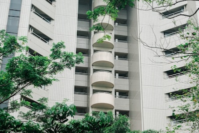 A high-rise apartment building with a modern facade featuring tiled exteriors and numerous balconies. The structure is partially obscured by lush, green trees and foliage in the foreground, adding a natural element to the urban scene.
