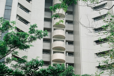 A high-rise apartment building with a modern facade featuring tiled exteriors and numerous balconies. The structure is partially obscured by lush, green trees and foliage in the foreground, adding a natural element to the urban scene.