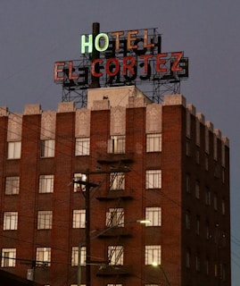 A multi-story brick building with a vintage style, featuring an illuminated neon sign that reads 'HOTEL EL CORTEZ'. The facade includes a detailed cornice and multiple windows, with an exterior fire escape visible. Overhead utility lines and a streetlamp are present in the foreground against a dusky sky.