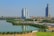 A cityscape featuring high-rise buildings near a water body with reflections on the surface. The foreground includes greenery and parked vehicles, and the urban skyline stretches out in the background under a clear blue sky.