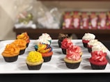 A variety of cupcakes with colorful frosting arranged neatly on a white rectangular platter. Each cupcake features a unique design, including orange, yellow, red, white, chocolate, and rainbow swirl frostings. The cupcakes are set against a blurred background with shelves containing packaged items.