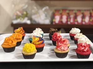 A variety of cupcakes with colorful frosting arranged neatly on a white rectangular platter. Each cupcake features a unique design, including orange, yellow, red, white, chocolate, and rainbow swirl frostings. The cupcakes are set against a blurred background with shelves containing packaged items.