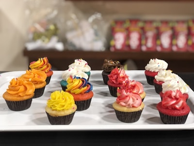 A variety of cupcakes with colorful frosting arranged neatly on a white rectangular platter. Each cupcake features a unique design, including orange, yellow, red, white, chocolate, and rainbow swirl frostings. The cupcakes are set against a blurred background with shelves containing packaged items.