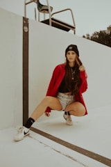 A person wearing a red jacket, black beanie, and white sneakers is crouching on the edge of an empty swimming pool. The individual has long hair and is dressed in casual attire, including denim shorts and a black shirt. A lifeguard chair is positioned in the background against a light sky, with trees visible nearby.