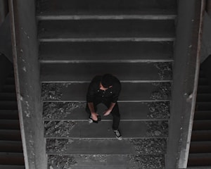 A person dressed in dark clothing sits thoughtfully on a dusty staircase surrounded by debris. The setting appears to be dimly lit and slightly industrial or abandoned, with broken bits scattered along the stairs.
