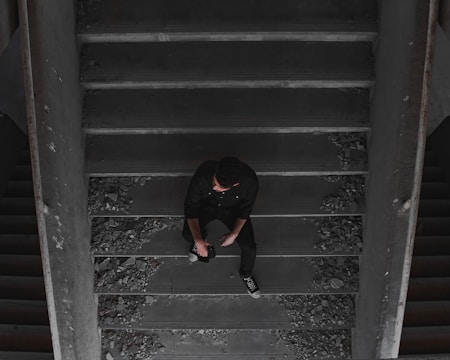 A person dressed in dark clothing sits thoughtfully on a dusty staircase surrounded by debris. The setting appears to be dimly lit and slightly industrial or abandoned, with broken bits scattered along the stairs.