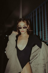 A person with glasses is wearing a black outfit with a deep neckline and a beige jacket. The individual, who has light brown hair styled in a middle part, is posing confidently in an urban setting with metal railings visible in the background.