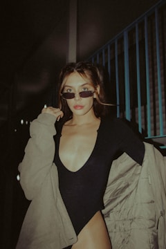 A person with glasses is wearing a black outfit with a deep neckline and a beige jacket. The individual, who has light brown hair styled in a middle part, is posing confidently in an urban setting with metal railings visible in the background.