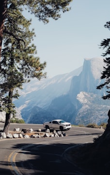 A white pickup truck is parked on a winding mountain road, surrounded by tall pine trees. In the background, there are expansive views of rocky mountains and cliffs under a clear blue sky.