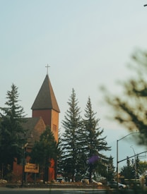 A red brick church with a pointed tower topped with a cross is situated among tall evergreen trees. The foreground includes a street with a traffic light and signage, contributing to a serene, small-town atmosphere.