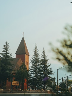 A red brick church with a pointed tower topped with a cross is situated among tall evergreen trees. The foreground includes a street with a traffic light and signage, contributing to a serene, small-town atmosphere.