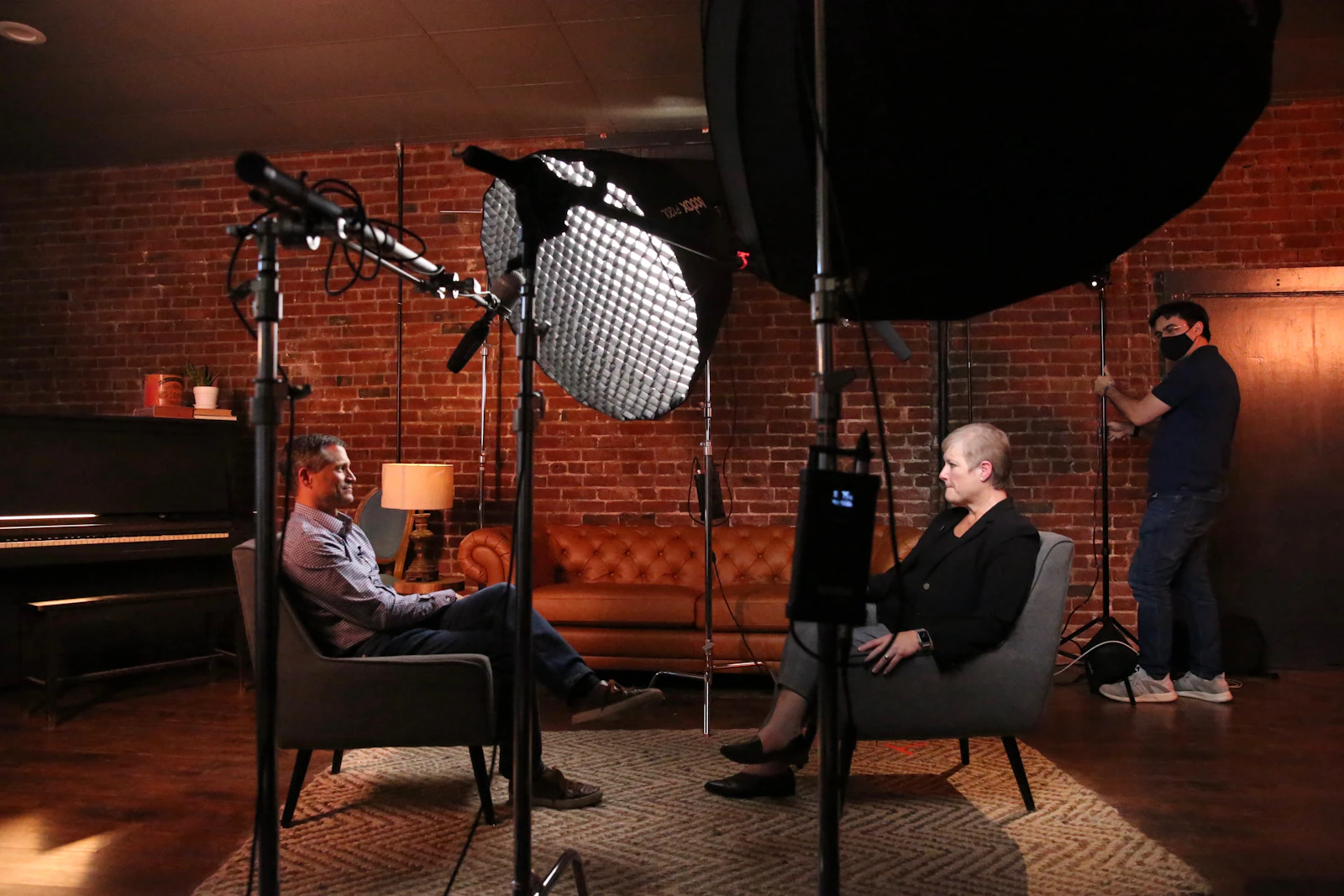 A two-person sit-down interview lit by a large softbox in a brick room, with a boom microphone overhead