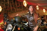 A woman in a brown leather jacket is posing confidently beside a motorcycle. She is holding a black helmet with one hand and sitting on the bike. The background features a warmly lit, elegant indoor setting with chandeliers and draped ceilings.