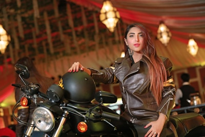 A woman in a brown leather jacket is posing confidently beside a motorcycle. She is holding a black helmet with one hand and sitting on the bike. The background features a warmly lit, elegant indoor setting with chandeliers and draped ceilings.