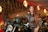 A woman in a brown leather jacket is posing confidently beside a motorcycle. She is holding a black helmet with one hand and sitting on the bike. The background features a warmly lit, elegant indoor setting with chandeliers and draped ceilings.