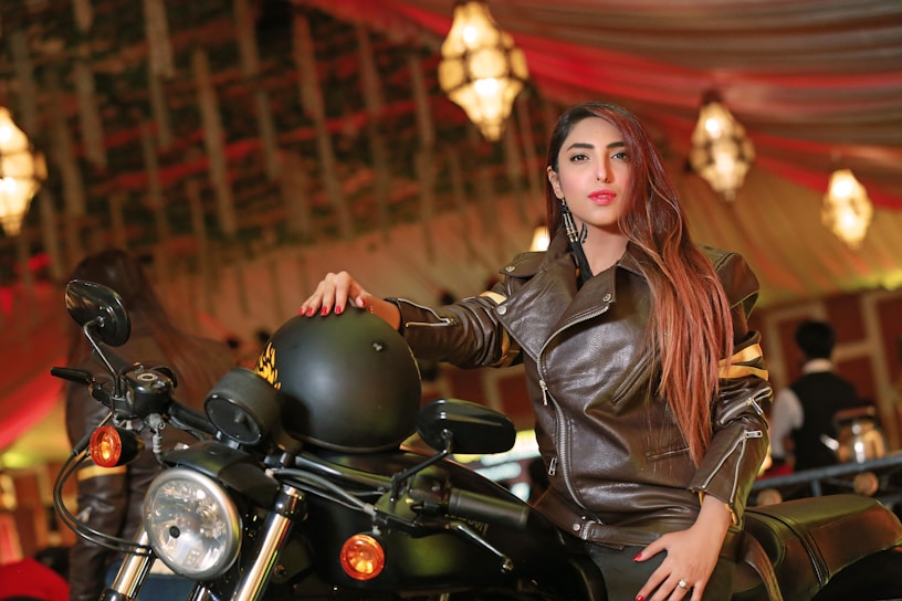 A woman in a brown leather jacket is posing confidently beside a motorcycle. She is holding a black helmet with one hand and sitting on the bike. The background features a warmly lit, elegant indoor setting with chandeliers and draped ceilings.