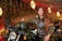 A woman in a brown leather jacket is posing confidently beside a motorcycle. She is holding a black helmet with one hand and sitting on the bike. The background features a warmly lit, elegant indoor setting with chandeliers and draped ceilings.