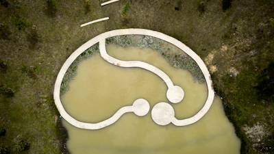 Aerial view of a modern, abstract landscape design featuring a concrete pathway over a murky pond. The pathway forms an interconnected loop with circular platforms. Surrounding the water are grassy areas with scattered trees and patches of shrubbery along the water's edge.