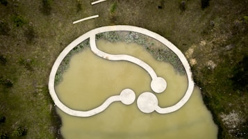 Aerial view of a modern, abstract landscape design featuring a concrete pathway over a murky pond. The pathway forms an interconnected loop with circular platforms. Surrounding the water are grassy areas with scattered trees and patches of shrubbery along the water's edge.