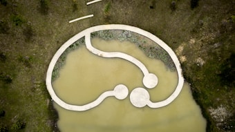 Aerial view of a modern, abstract landscape design featuring a concrete pathway over a murky pond. The pathway forms an interconnected loop with circular platforms. Surrounding the water are grassy areas with scattered trees and patches of shrubbery along the water's edge.