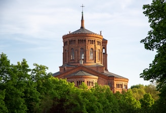 A large, historic brick building with a circular tower and a cross on top, surrounded by lush green trees under a clear blue sky.