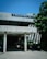 A concrete building with a sign labeled '3 Metallurgy Depart' is partially shaded by a large tree with green foliage. The building has columns supporting a roof over a small entrance area where several motorcycles are parked. The sky is clear and blue, contributing to a sunny atmosphere.