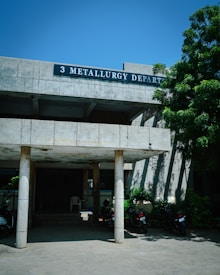 A concrete building with a sign labeled '3 Metallurgy Depart' is partially shaded by a large tree with green foliage. The building has columns supporting a roof over a small entrance area where several motorcycles are parked. The sky is clear and blue, contributing to a sunny atmosphere.