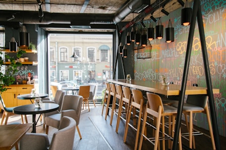 A stylish cafe interior features modern decor with a long wooden bar counter and light wooden chairs. The wall has colorful graffiti art and is adorned with multiple pendant lights. A cozy seating area has round tables, soft chairs, and small vases with flowers. Large windows let in ample natural light, offering a view of the building across the street.