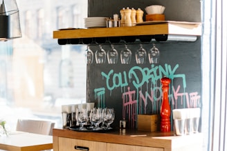 A countertop setup inside a cafe or restaurant, featuring hanging wine glasses, pepper grinders, and stacked plates. A blue and pink graffiti message on the wall reads 'You Drink I Work'.