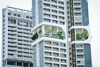 Modern high-rise residential buildings with a unique architectural design featuring prominent balconies and integrated greenery. The structures have a geometric pattern with large windows and white, sleek facades.