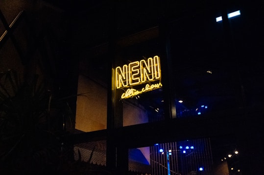 A bright yellow neon sign with the text 'NENI' is visible against a dark background. The sign is enclosed in a window frame, and below it, there is a cursive script that is less legible. Some blue lights are subtly glowing in the background, likely reflecting an indoor atmosphere.