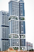 A modern high-rise building with a distinctive architectural design, featuring large, open sections and integrated greenery. The building's facade is characterized by a combination of blue and white colors, incorporating numerous windows and geometric shapes. Smaller, traditional houses with red-tiled roofs are visible at the base, creating a contrast with the towering structure above.