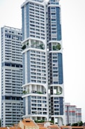 A modern high-rise building with a distinctive architectural design, featuring large, open sections and integrated greenery. The building's facade is characterized by a combination of blue and white colors, incorporating numerous windows and geometric shapes. Smaller, traditional houses with red-tiled roofs are visible at the base, creating a contrast with the towering structure above.