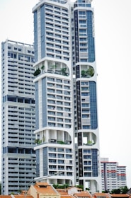 A modern high-rise building with a distinctive architectural design, featuring large, open sections and integrated greenery. The building's facade is characterized by a combination of blue and white colors, incorporating numerous windows and geometric shapes. Smaller, traditional houses with red-tiled roofs are visible at the base, creating a contrast with the towering structure above.