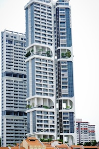 A modern high-rise building with a distinctive architectural design, featuring large, open sections and integrated greenery. The building's facade is characterized by a combination of blue and white colors, incorporating numerous windows and geometric shapes. Smaller, traditional houses with red-tiled roofs are visible at the base, creating a contrast with the towering structure above.