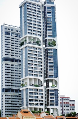 A modern high-rise building with a distinctive architectural design, featuring large, open sections and integrated greenery. The building's facade is characterized by a combination of blue and white colors, incorporating numerous windows and geometric shapes. Smaller, traditional houses with red-tiled roofs are visible at the base, creating a contrast with the towering structure above.