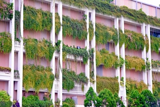 A building facade with pink walls features numerous vertical green gardens. Lush plants and vines cascade down from multiple levels, integrated into the structure's design, creating a living wall effect. Trees line the base, enhancing the natural aesthetics.