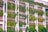 A building facade with pink walls features numerous vertical green gardens. Lush plants and vines cascade down from multiple levels, integrated into the structure's design, creating a living wall effect. Trees line the base, enhancing the natural aesthetics.