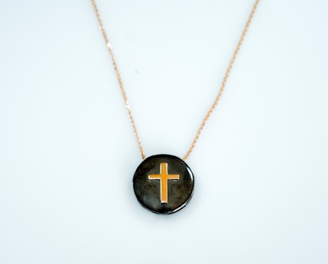 A pendant featuring a gold cross design set against a glossy dark background, suspended on a delicate gold chain.