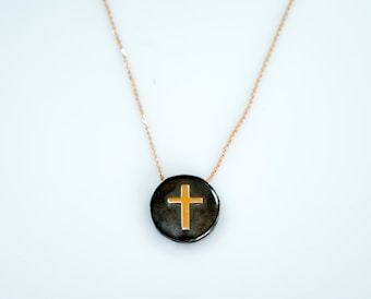 A pendant featuring a gold cross design set against a glossy dark background, suspended on a delicate gold chain.