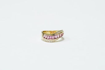A gold ring featuring a central band of pink rectangular gemstones, flanked by rows of small, clear stones. The ring appears well-crafted with a modern and elegant design.