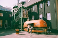 A retro-style orange food truck with the name 'Hungry Bro' on its side is stationed in an urban area. The truck is parked near a dark building that has multiple air conditioning units and a green metal fire escape on one side. A woman is standing near the service window of the food truck, likely ordering food.