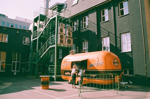 A retro-style orange food truck with the name 'Hungry Bro' on its side is stationed in an urban area. The truck is parked near a dark building that has multiple air conditioning units and a green metal fire escape on one side. A woman is standing near the service window of the food truck, likely ordering food.