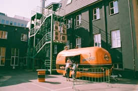 A retro-style orange food truck with the name 'Hungry Bro' on its side is stationed in an urban area. The truck is parked near a dark building that has multiple air conditioning units and a green metal fire escape on one side. A woman is standing near the service window of the food truck, likely ordering food.