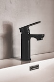 A modern black faucet is mounted on a white sink, set against a textured wall. The design is minimalist, featuring clean lines and a matte finish. The shadow of the faucet is cast on the wall, adding depth to the composition.