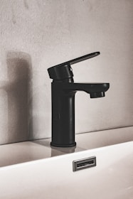 A modern black faucet is mounted on a white sink, set against a textured wall. The design is minimalist, featuring clean lines and a matte finish. The shadow of the faucet is cast on the wall, adding depth to the composition.