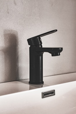 A modern black faucet is mounted on a white sink, set against a textured wall. The design is minimalist, featuring clean lines and a matte finish. The shadow of the faucet is cast on the wall, adding depth to the composition.