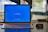 A laptop on a desk is displaying a blue screen with an update notification. The screen shows a progress indicator for Windows updates at 27%, along with text advising not to turn off the PC. A mouse is placed next to the laptop on the desk.